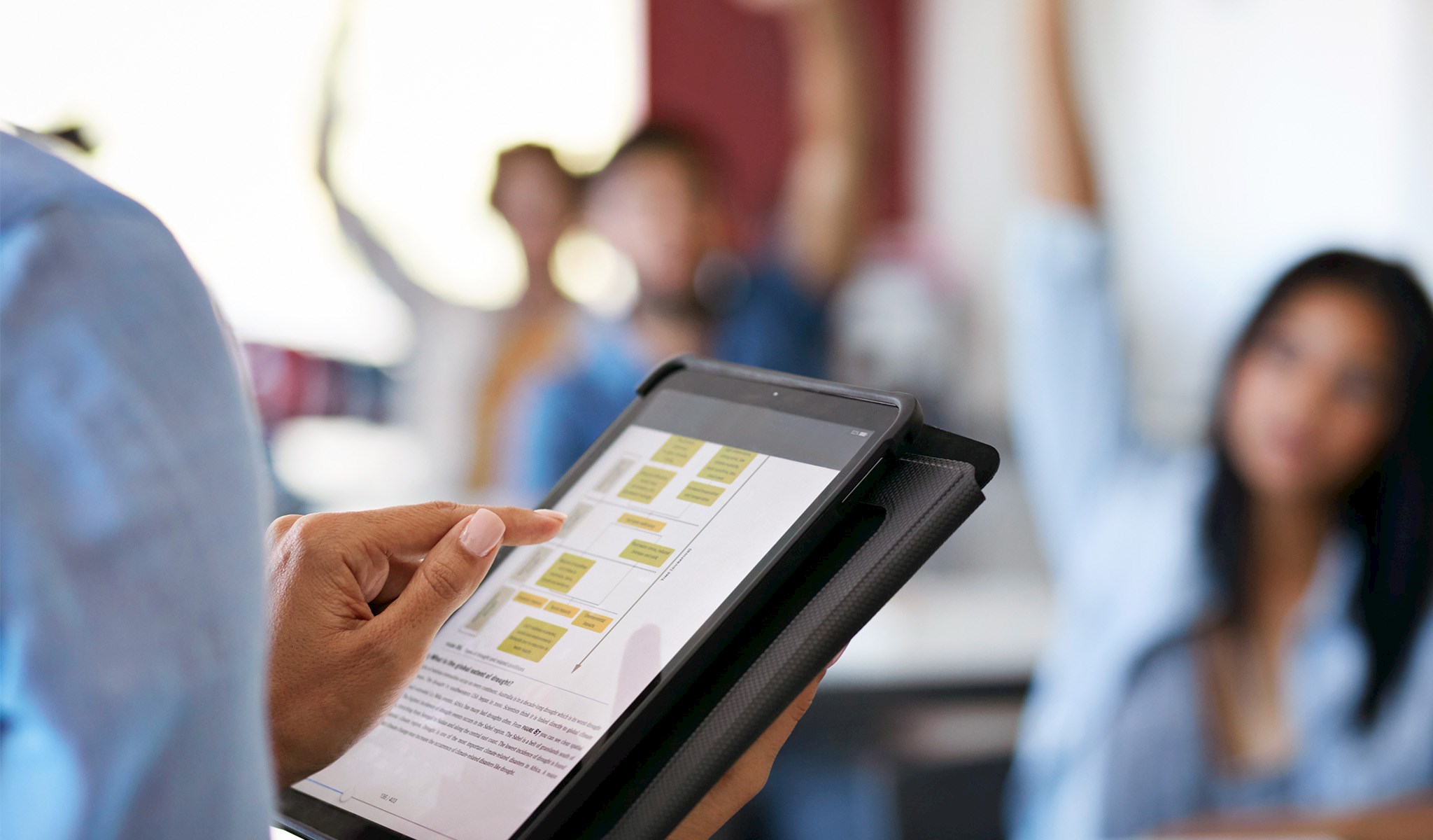 Close up of a teacher holding a tablet in front of a classroom of students